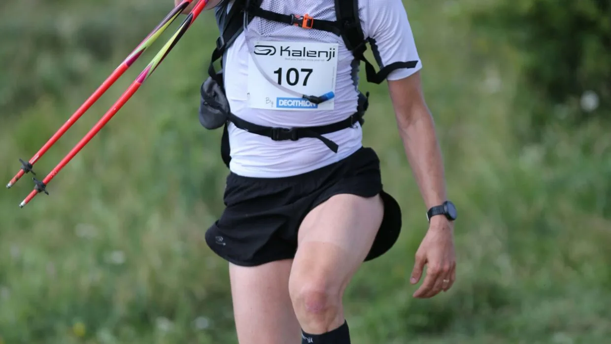 Corredor caminhando na subida de trail running em trilha íngreme com bastões nas mãos