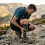 Corredor com câimbra no trail agachado em trilha rochosa segurando a panturrilha durante prova de montanha