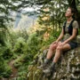 Trail running faz bem para a saúde mental? 6 Trail running saúde mental: mulher descansando em pedra no topo de trilha com natureza ao fundo e luz suave da manhã