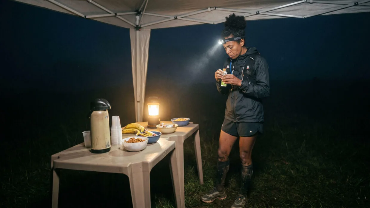 Nutrição para ultra trail de inverno em altitude: o que muda quando o frio e a hipóxia entram em cena 10 Nutrição ultra trail inverno: corredora se alimentando em posto de abastecimento noturno em campo de altitude